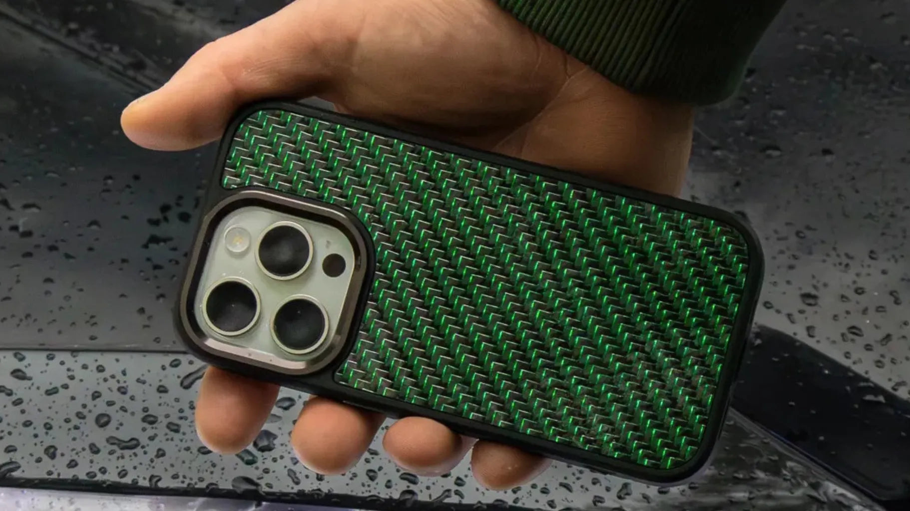 Close-up of a green carbon fiber phone case held in hand with water droplets on a car hood in the background.