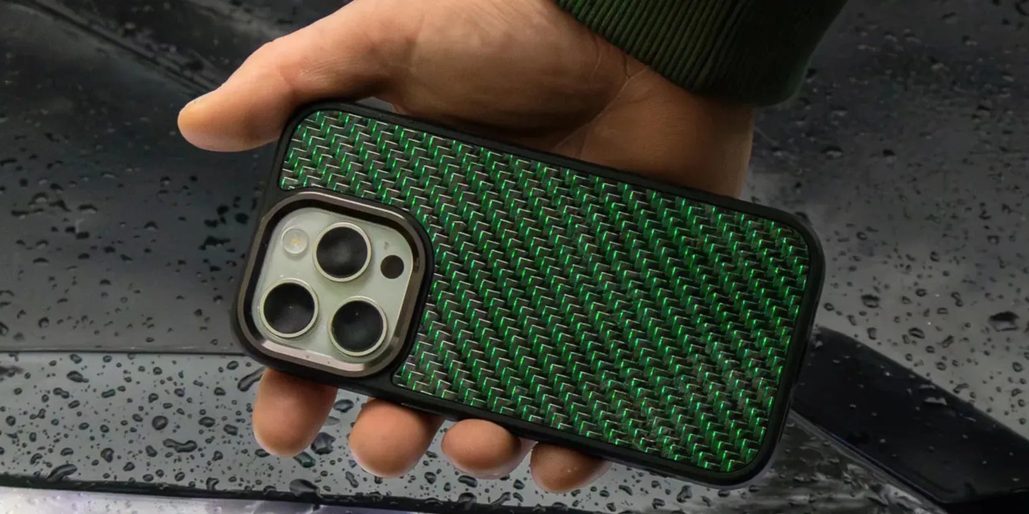 Close-up of a green carbon fiber phone case held in hand with water droplets on a car hood in the background.