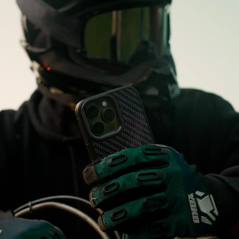 Motocross rider holding a carbon fiber phone case while sitting on a dirt bike, helmet on, ready to ride.